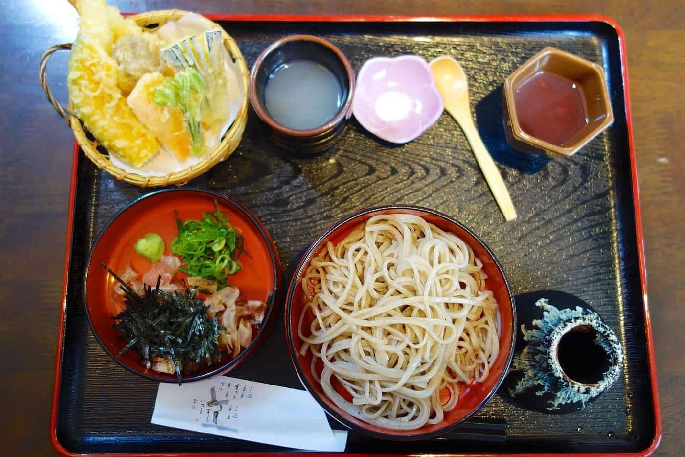 A tray with cold soba noodles, tempura, dipping sauce and small side dishes