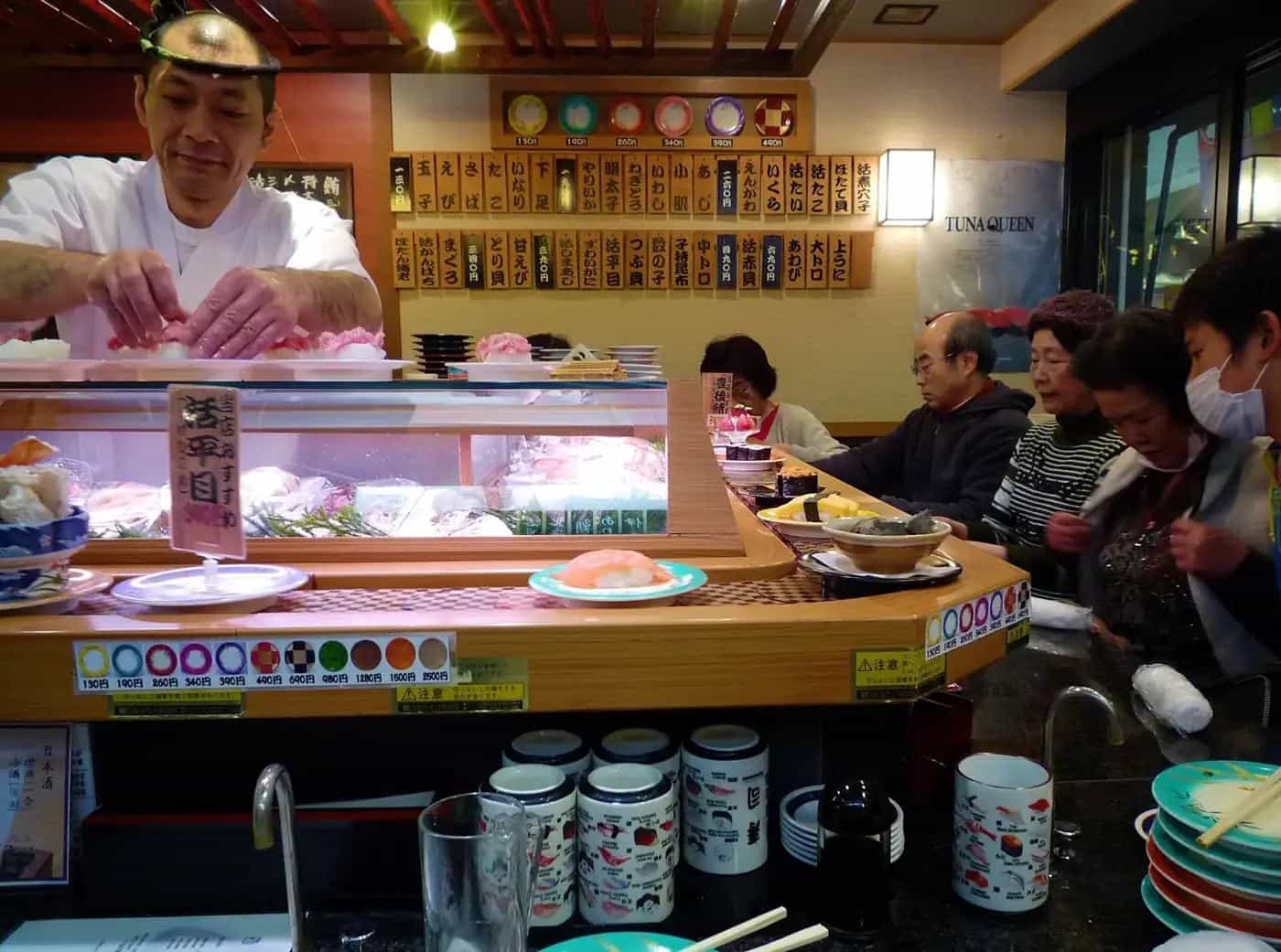 A sushi chef preparing nigiri behind a conveyor-belt sushi counter while diners eat