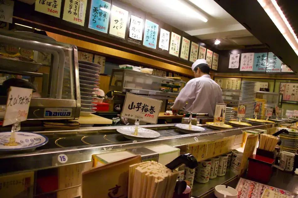 A chef working behind the conveyor belt at a small sushi bar, with handwritten menu signs on the wall